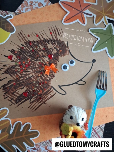A fork painted hedgehog craft made with brown paint on paper, decorated with googly eyes, red star confetti, and an orange bow, is surrounded by leaf cutouts and a small hedgehog figurine.