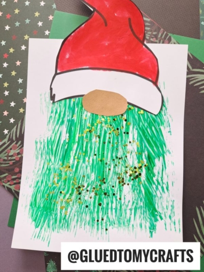 A handmade Santa gnome craft featuring a red hat, oval beige nose, and a fork painted Grinch-green beard with gold and black glitter, displayed on white paper over holiday-themed backgrounds. @GLUEDTOMYCRAFTS is written at the bottom.