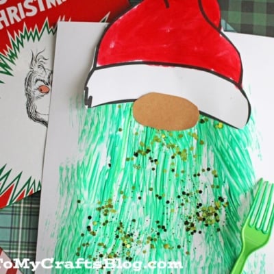 A children’s craft project features a fork painted Grinch-inspired gnome with a red Santa hat and a textured green beard, displayed on white paper beside a green plastic fork. “How the Grinch Stole Christmas!” book sits in the background.