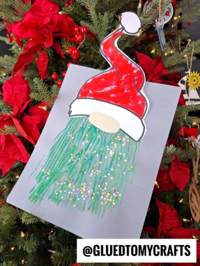 A paper craft of a gnome with a red Santa hat and a glittery green, fork painted Grinch-inspired beard is displayed on a gray sheet, placed against a decorated Christmas tree with red poinsettias. @GLUEDTOMYCRAFTS is labeled at the bottom.