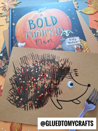 A childrens book titled A Bold Pumpkin Plan lies behind a fork painted hedgehog craft made from brown paint, fall leaf confetti, a googly eye, and a fork on brown paper. The handle of the fork is visible nearby.