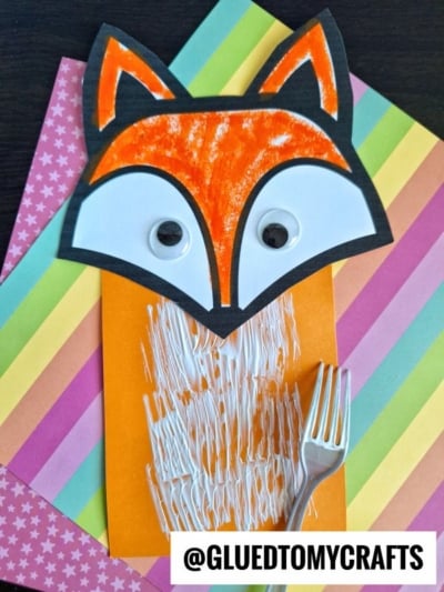 A fork painted fox craft with googly eyes on colorful paper; its orange body features white paint textured by a plastic fork. @GLUEDTOMYCRAFTS is written at the bottom.