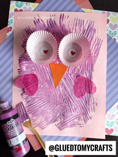 A fork painted owl craft featuring cupcake liner eyes, paper heart wings, a triangle orange beak, and textured purple paint strokes on pink paper, all displayed on patterned paper backgrounds.