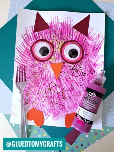 A fork painted owl craft featuring textured pink paint strokes, large googly eyes, and paper shapes for the ears, wings, and beak. A plastic fork, gold glitter, and a bottle of pink paint are arranged on a pastel background.
