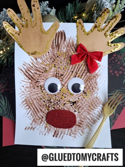 A fork painted reindeer craft made from brown paint strokes, large googly eyes, a red glittery mouth, a red bow, and handprint antlers with gold glitter is shown on white paper next to a plastic fork.