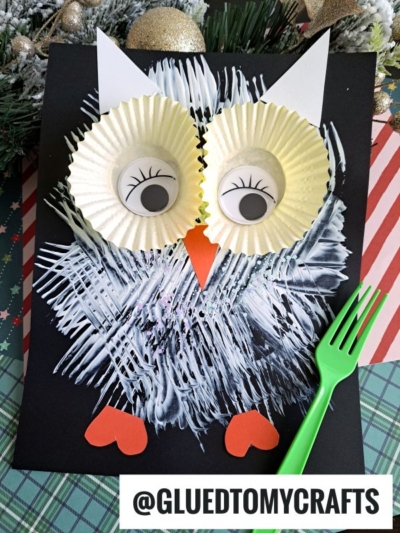 A fork painted owl with cupcake liner eyes, drawn eyelashes, a paper beak and feet, and white streaks on black paper. A green plastic fork is nearby, with Christmas decorations in the background.