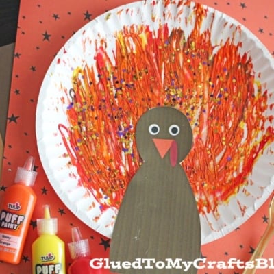 A fork painted turkey sits on a star-patterned background, featuring a paper plate with orange, red, and yellow feathers and a brown paper body. A gold fork and colorful paint bottles add to the creative scene.