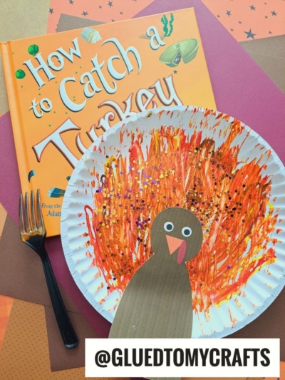 A fork painted turkey craft with orange and red paint and glitter sits next to the book How to Catch a Turkey, a black fork, and colored paper. The turkey’s body is made from brown paper, with handle @GLUEDTOMYCRAFTS at the bottom.