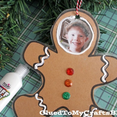 A Gingerbread Man Photo Ornament made from brown paper with a child’s photo as the face, decorated with white squiggles and glittery buttons, lies on plaid fabric near a Christmas tree.
