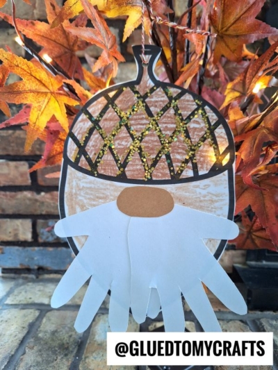 A handmade acorn gnome craft with a paper acorn hat, white handprint beard, and brown oval nose, hanging in front of autumn leaves. The photo includes the text @GLUEDTOMYCRAFTS at the bottom.