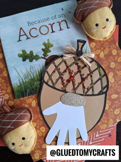 A fall-themed craft scene with the book Because of an Acorn, two stuffed acorn plushies, a charming acorn gnome, a paper acorn craft featuring a white handprint, and a @GLUEDTOMYCRAFTS label on autumn-patterned paper.