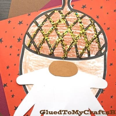 This handmade acorn gnome craft features a large acorn cap with gold glitter, a white handprint beard, and a brown oval nose. Set against red paper with black stars, this charming acorn gnome makes a whimsical fall decoration.
