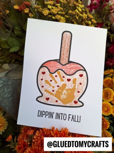 A handmade fall card features a handprint caramel apple outline with a child’s golden handprint inside, red hearts, and the text “DIPPIN INTO FALL!” amid autumn flowers. The handle @GLUEDTOMYCRAFTS appears at the bottom.
