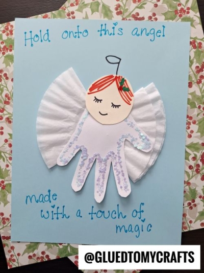 A handmade angel craft on blue paper, made with a child’s handprint, a coffee filter as wings, a paper face, and glitter—perfect for christmas keepsakes. Text reads: “Hold onto this angel made with a touch of magic.”.