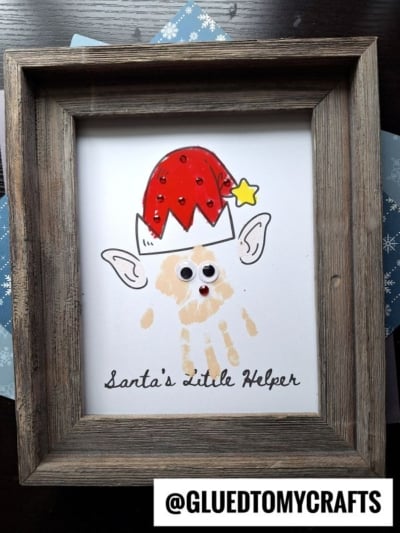 A framed craft features a handprint elf turned into a Christmas character with googly eyes, pointed ears, and a red hat topped with a yellow star. The text reads “Santa’s Little Helper,” set against blue snowflake paper in the background.