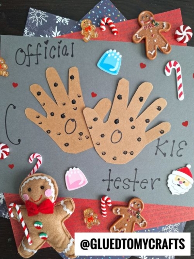 A craft project features two brown paper handprints decorated as chocolate chip cookies on gray paper, surrounded by festive stickers, candy canes, and gingerbread figures—perfect for christmas keepsakes. The text reads Official Cookie Tester.
