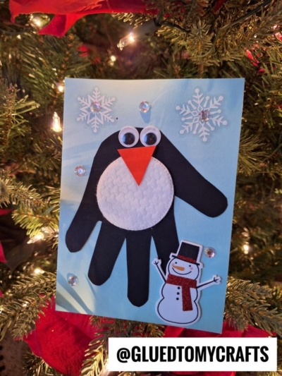 A handmade penguin card hangs on a Christmas tree—crafted from a black handprint with googly eyes, an orange beak, and textured white belly. Snowflakes, gems, and a snowman sticker make it perfect for christmas keepsakes.