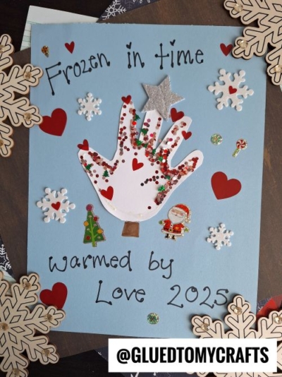 A blue card decorated with a red and white handprint, glitter, snowflake and heart stickers, and the words Frozen in time warmed by love 2025—perfect for christmas keepsakes. The Instagram handle @GLUEDTOMYCRAFTS appears at the bottom.