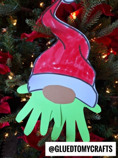 A Christmas tree decorated with a handmade Grinch Gnome ornament featuring a red Santa hat, a brown oval nose, and a green beard made from cut-out hand shapes. Text at the bottom reads @GLUEDTOMYCRAFTS.