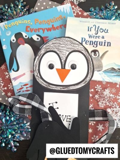 A handprint penguin craft made from paper with googly eyes and an orange beak is displayed on top of a snowflake-patterned background, alongside two children’s books about penguins.