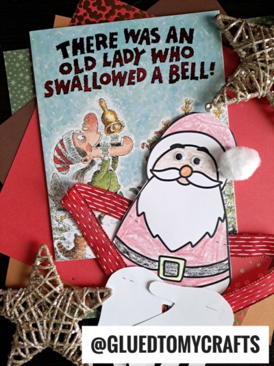 A children’s book titled “There Was An Old Lady Who Swallowed a Bell!” is shown with a handprint Santa hug craft, glittery star ornaments, red ribbon, colored paper, and the text @GLUEDTOMYCRAFTS in the foreground.