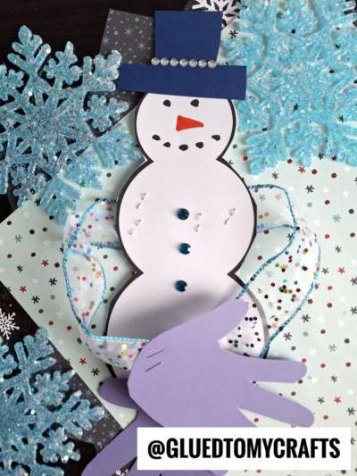 A handmade snowman craft with a blue hat, rhinestone buttons, and a purple handprint scarf—like a warm snowman hug—surrounded by glittery snowflakes and star-patterned paper. @GLUEDTOMYCRAFTS is written at the bottom.