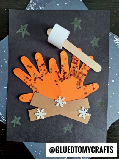 A craft project showing two orange paper handprints as a campfire on black paper, with gold glitter as flames, brown paper logs, white snowflake stickers, and a marshmallow on a stick—perfect for making festive Christmas keepsakes.