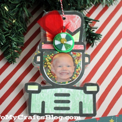 A handmade nutcracker photo ornament shaped like a toy soldier features a child’s photo in the face area, decorated with glitter, a red feather, and a green gem. It hangs from a Christmas tree with candy cane-striped paper beneath.