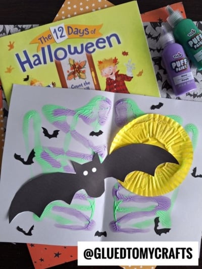 A Halloween craft scene features a black paint splat bat on white paper, accented with purple and green squiggles, small bat cutouts, a yellow moon, and the 12 Days of Halloween book. Text: @GLUEDTOMYCRAFTS.