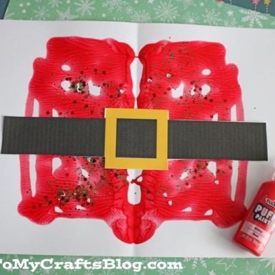 A festive paint splat Santa belly features red paint, gold glitter, a black paper belt, and yellow buckle on white paper. Nearby sit red paint and green festive paper.