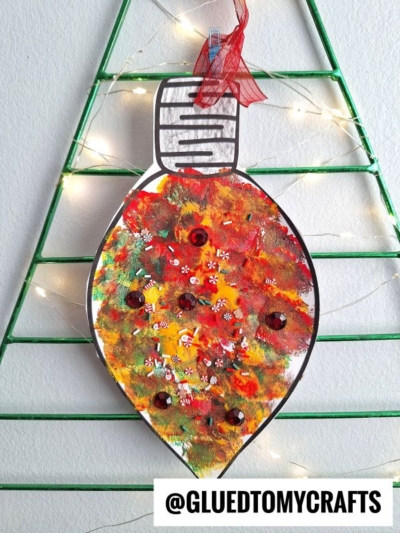 A colorful, hand-painted pom pom stamped ornament with sequins and glitter hangs on a green wire tree frame decorated with string lights. The handle @GLUEDTOMYCRAFTS is at the bottom.