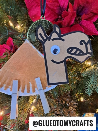 A handmade paper plate camel Christmas ornament with a paper head, painted body, clothespin legs, and a googly eye hangs on a decorated tree. A medal and red poinsettia flowers are also visible. @GLUEDTOMYCRAFTS is labeled at the bottom.