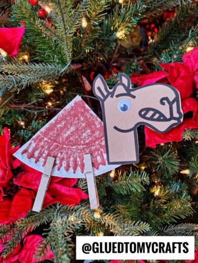 A handmade paper plate camel ornament with a googly eye and red painted blanket, crafted from paper, clothespins, and a cupcake liner, hangs on a Christmas tree adorned with red poinsettias and glowing lights. The handle @GLUEDTOMYCRAFTS is at the bottom.