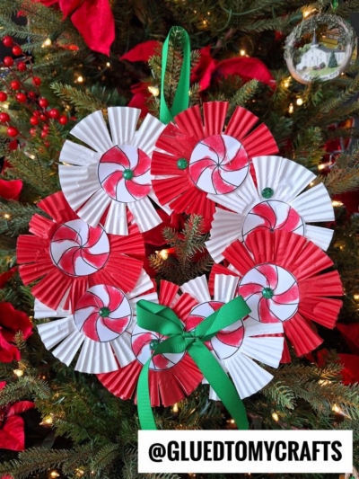 A Peppermint Candy Wreath made of red and white paper rosettes, decorated with a green ribbon bow, hangs on a Christmas tree. Red berries and ornaments add festive charm in the background.