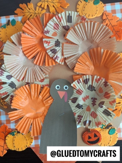A cupcake liner turkey craft made with orange and patterned liners for feathers, featuring a paper turkey body plus autumn leaf and pumpkin accents, all displayed on a checkered background.