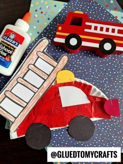 Children’s paper plate fire truck craft made with paper, tissue, and a plate, next to school glue, a toy fire truck, and blue polka dot paper. Text at bottom reads “@GLUEDTOMYCRAFTS.”.