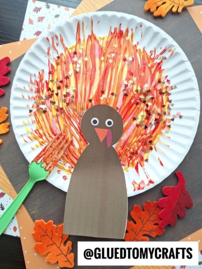 A fork painted turkey craft featuring a paper plate “feathered” with fall colors using a plastic fork, a brown paper turkey body, googly eyes, and surrounded by felt leaves. Text: @GLUEDTOMYCRAFTS.