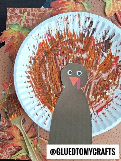 A fork painted turkey craft with a colorful feathered body made from brown, orange, and yellow paint, a paper cut-out turkey head and body, plus decorative autumn leaves, shown on a dotted background.