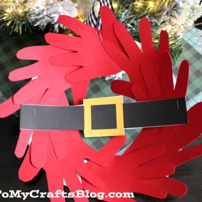 A handmade handprint Santa wreath crafted from red paper handprints, decorated with a black paper belt and yellow buckle to resemble Santa’s suit.