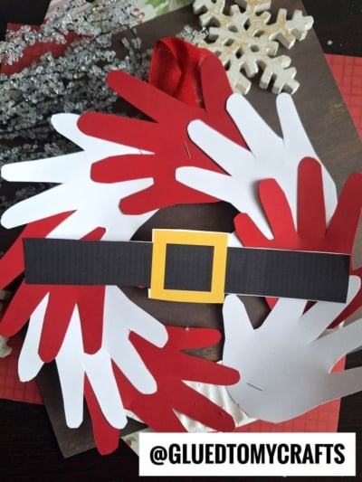 A handprint Santa wreath made from red and white paper cutouts, decorated with a black paper belt and yellow buckle, is displayed on a table. The text @GLUEDTOMYCRAFTS appears in the corner.