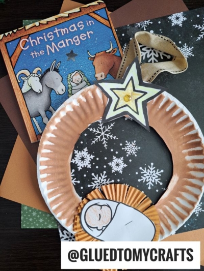 A handmade Paper Plate Nativity Wreath craft, created with gold paper and a star, is displayed beside the children’s book Christmas in the Manger and colorful construction paper. @GLUEDTOMYCRAFTS.