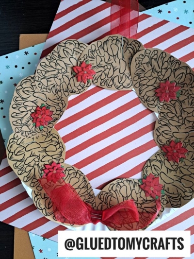 A handmade pinecone wreath crafted from paper handprints with red flower embellishments and a red ribbon bow, displayed on a red and white striped background. @GLUEDTOMYCRAFTS is written at the bottom.