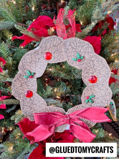 A handmade pinecone wreath with black squiggly lines, red gems, and holly stickers hangs on a Christmas tree. The wreath is decorated with a large red ribbon bow. The handle @GLUEDTOMYCRAFTS is at the bottom.