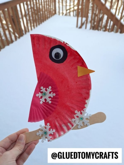 A hand holds a paper plate cardinal craft, featuring a painted plate, googly eye, orange beak, snowflake stickers, and wooden stick handle. A snowy deck is in the background. @GLUEDTOMYCRAFTS is labeled at the bottom.