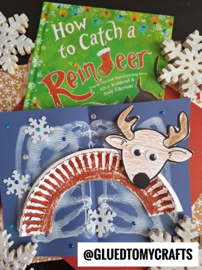 A children’s book titled How to Catch a Reindeer is pictured above a handmade reindeer Snow Craft Idea For Kids, featuring a brown-painted paper plate, paper snowflakes, blue gems, and the handle @GLUEDTOMYCRAFTS.