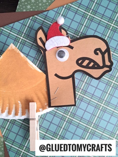 A handmade paper plate camel craft featuring a googly eye and a tiny red Santa hat, made with paper and a clothespin. It’s set on plaid and brown backgrounds. Text reads “@GLUETOMYCRAFTS.”.