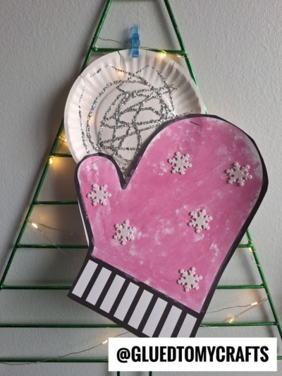 A pink paper mitten decorated with white snowflakes is displayed in front of a paper plate snowball with silver glitter swirls. Both are clipped to a green wire Christmas tree decorated with fairy lights. @GLUEDTOMYCRAFTS is at the bottom.