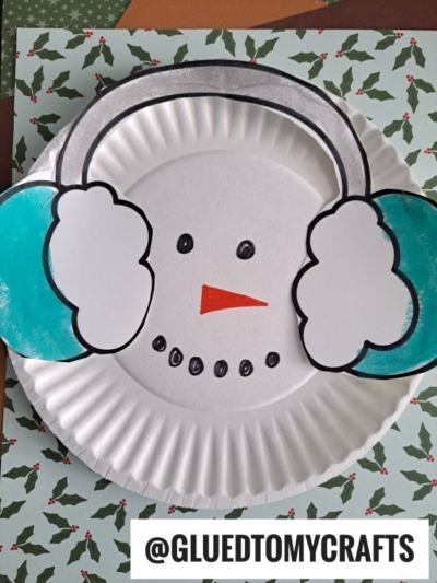 A paper plate snowman decorated as a snowman’s face with drawn eyes, mouth, an orange carrot nose, and blue earmuffs made from colored paper. @GLUEDTOMYCRAFTS is printed at the bottom.