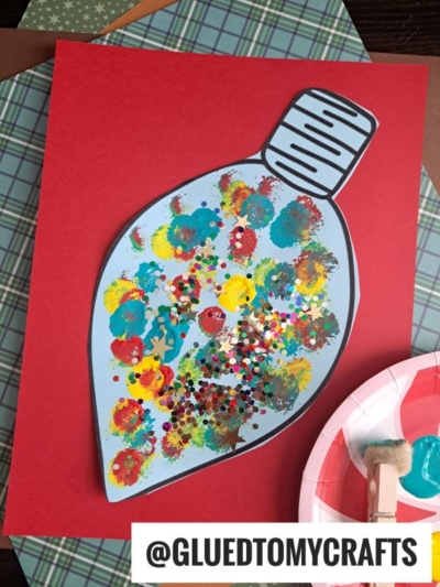 A large paper Christmas light bulb, decorated as a pom pom stamped ornament with colorful paint dots and glitter, sits on a red background. Nearby are craft supplies like patterned paper and a paint palette. @GLUEDTOMYCRAFTS is written at the bottom.