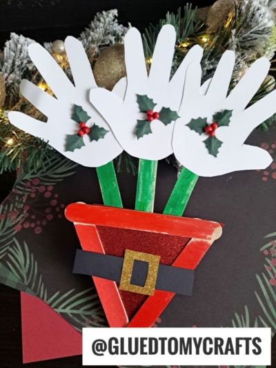 A festive craft shows a Christmas flower bouquet made from three white paper handprints decorated with holly leaves and berries, arranged in a red Santa-style flower pot of craft sticks, with pine garland behind and @GLUEDTOMYCRAFTS below.